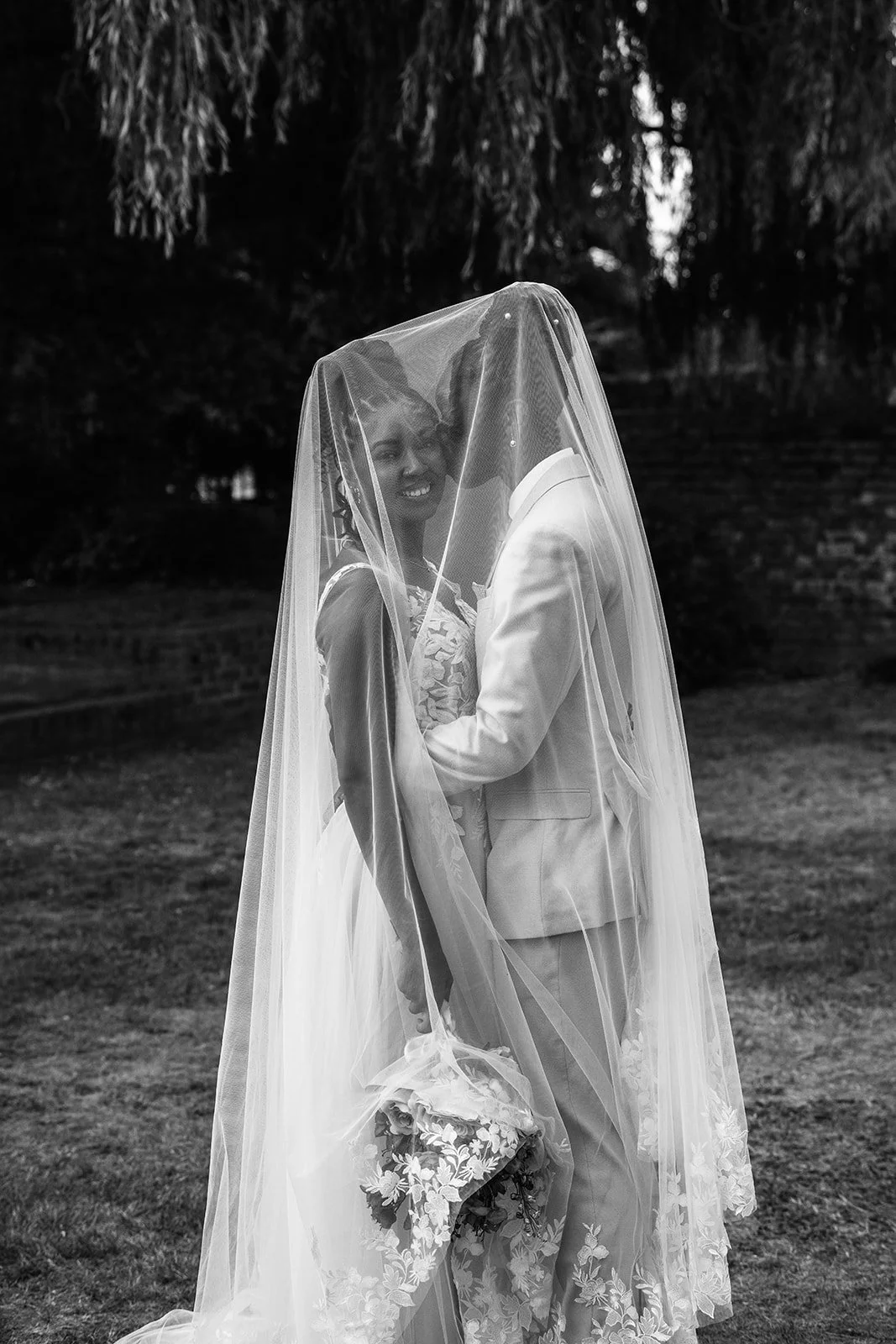 Black and white portrait of Kasey and Larisha standing close together beneath Larisha’s veil during their wedding portraits at Oaks Farm Weddings in Surrey.