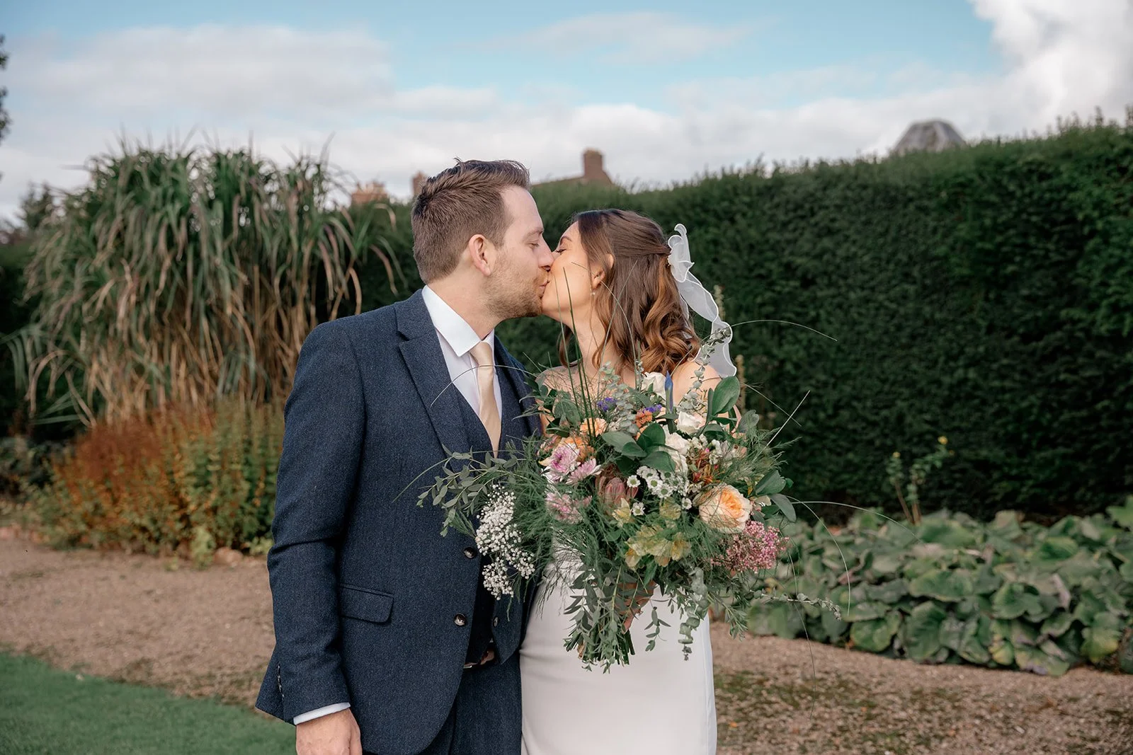 Lilly & Lloyd sharing a kiss in the gardens at Broadfield Court in Herefordshire, with the bride holding a colourful bouquet, photographed by Just James Weddings.