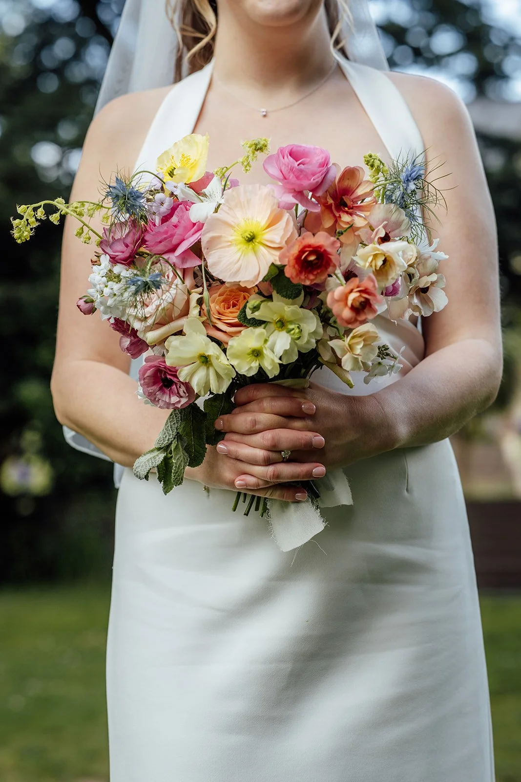 Close-up of Bryony’s colourful wedding bouquet at Insole Court in Cardiff, held against her white dress during Bryony & Max’s wedding, with pink, yellow, peach, white and blue flowers creating a fresh garden-inspired floral detail outdoors.