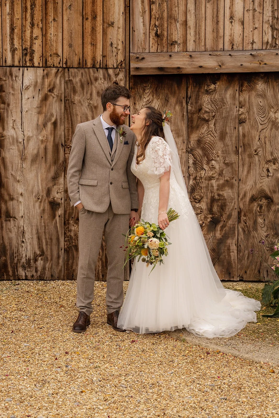 Maisie & Andrew standing together in front of rustic wooden barn doors at Stockbridge Farm Barn in Dorset, photographed by Just James Weddings for relaxed autumn wedding portraits.
