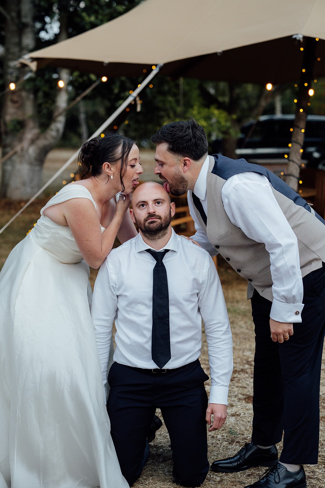 Harriet & Simon lean playfully towards a kneeling wedding guest during a fun candid moment outside the tipi at Dovecote Events in Oxfordshire, photographed by Just James Weddings.