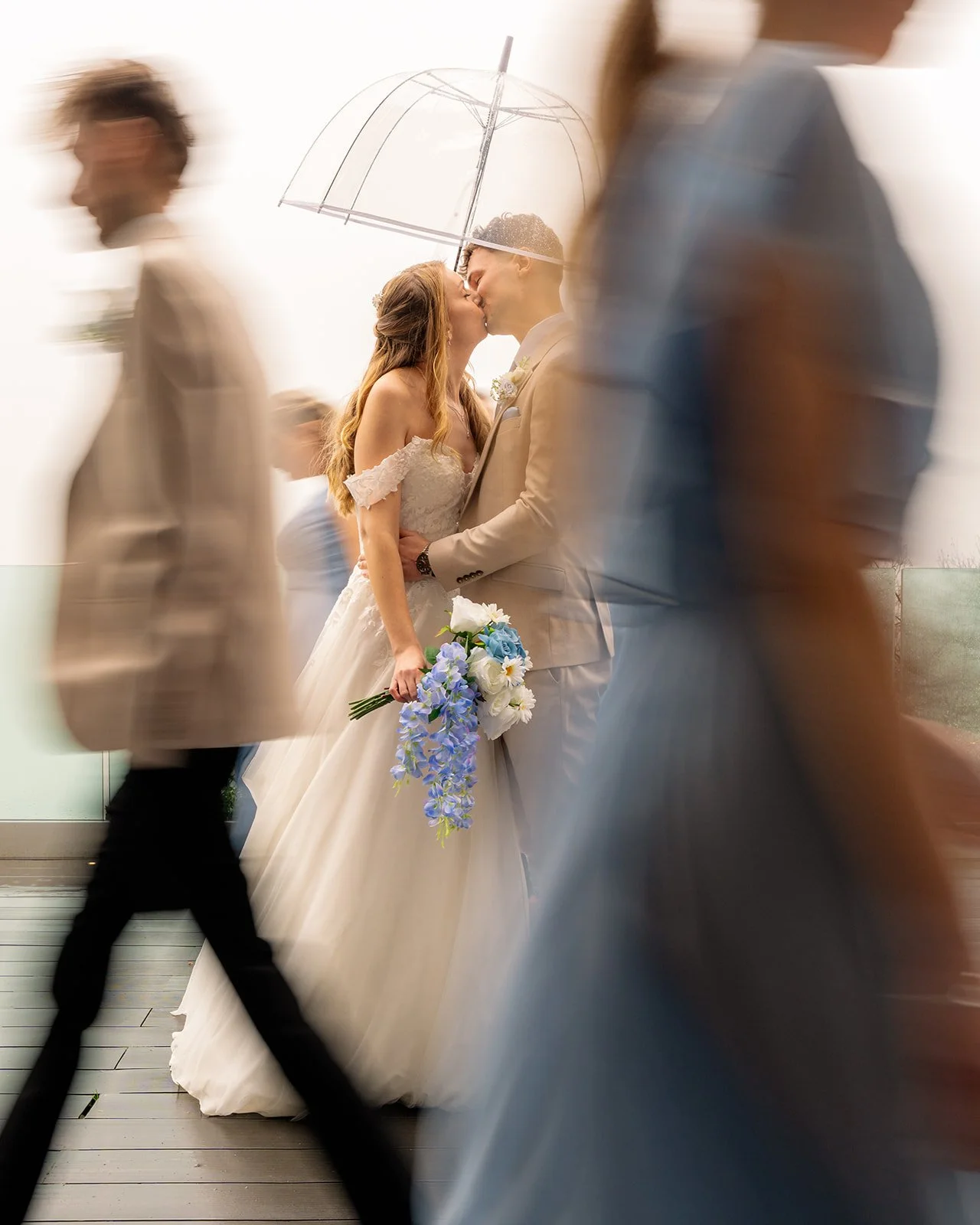 Hannah & Cameron kissing beneath a clear umbrella at Sandy Cove Hotel in North Devon, with guests passing in soft motion blur, photographed by Just James Weddings for romantic coastal wedding portraits with warm light and blue bouquet details.