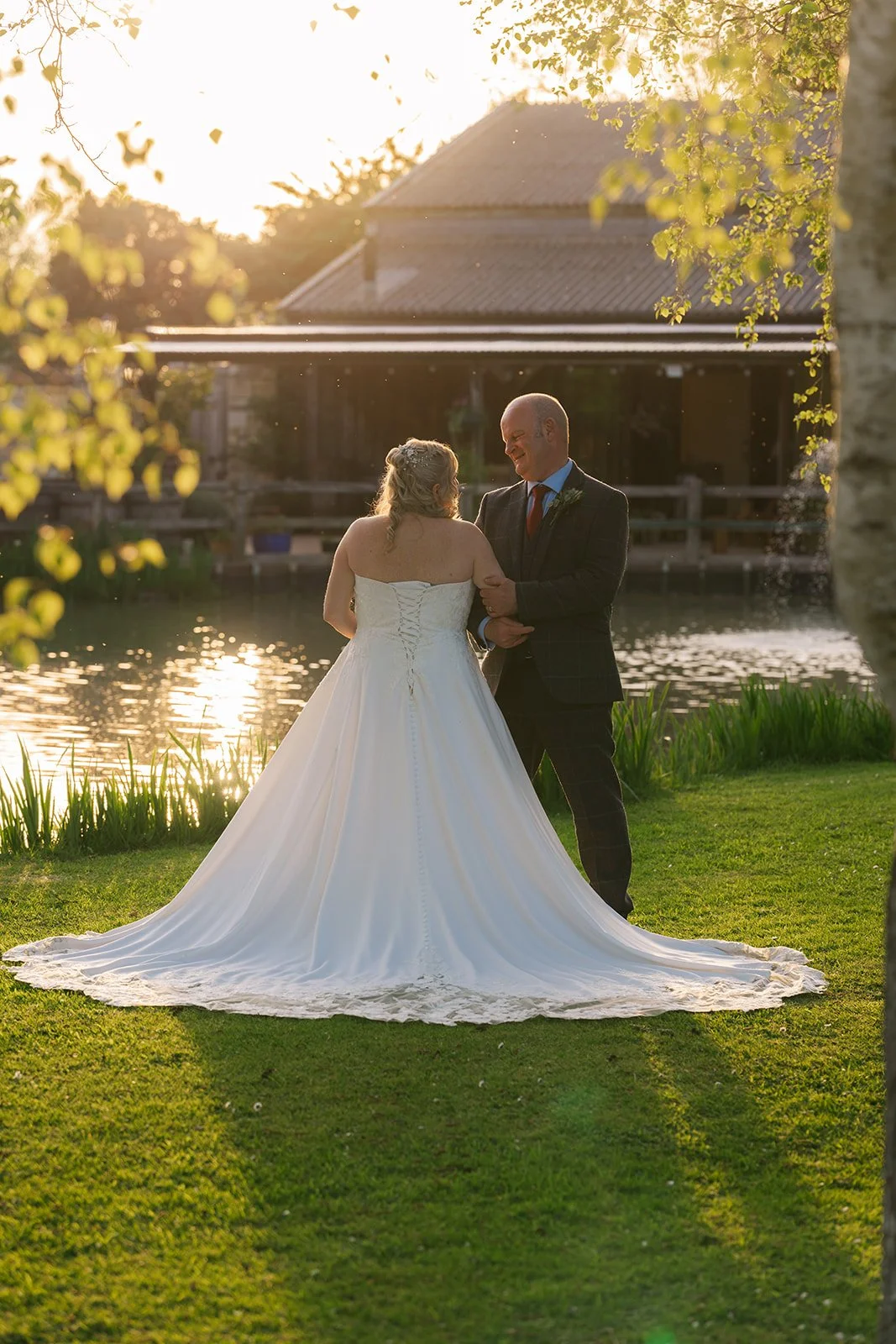 Debbie and Nigel stand together by the lake at Yarlington Barn in Somerset during golden hour, with warm light behind the barn, Debbie’s dress spread across the grass and a quiet look between them.