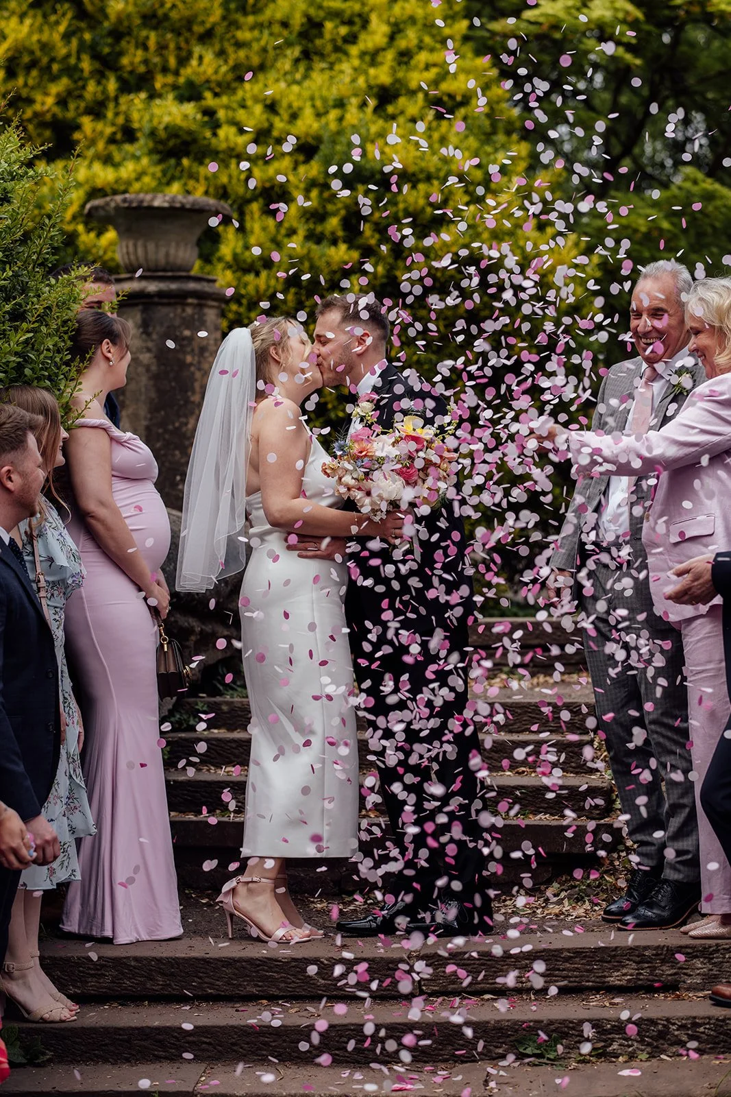 Bryony & Max kiss on the garden steps at Insole Court in Cardiff while guests throw pink and white confetti around them, capturing a joyful newlywed moment surrounded by family, friends and lush greenery after their ceremony outdoors there.
