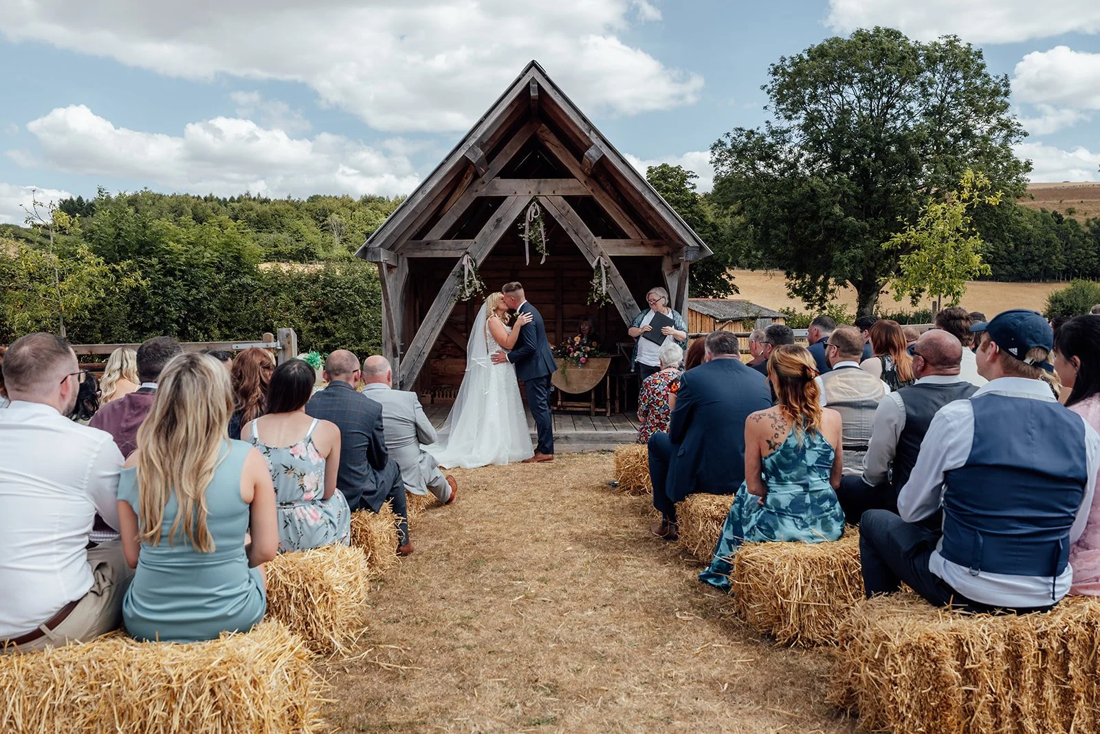 Elise & Tom kiss during their outdoor wedding ceremony at Bowerchalke Barn in Wiltshire, with guests seated on hay bales in the countryside, photographed by Just James Weddings.