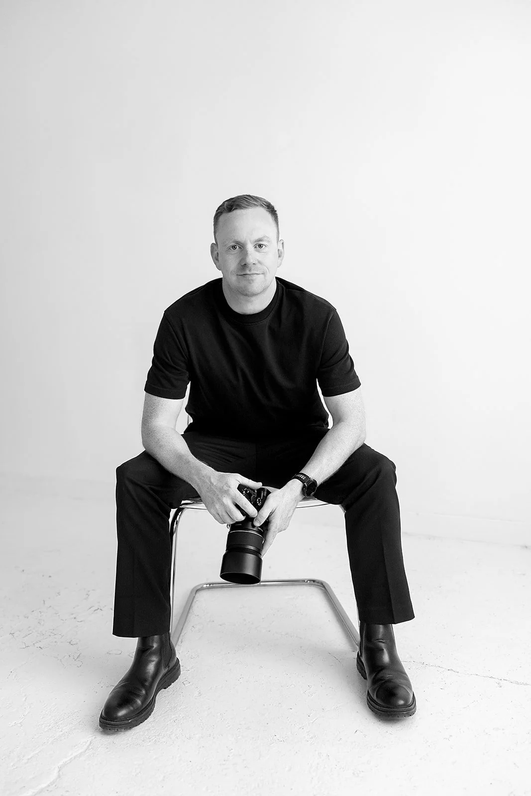 Black and white portrait of James from Just James Weddings sitting on a studio chair, holding a camera and looking towards the viewer against a clean white backdrop, for his relaxed Somerset wedding photography website about page and profile.