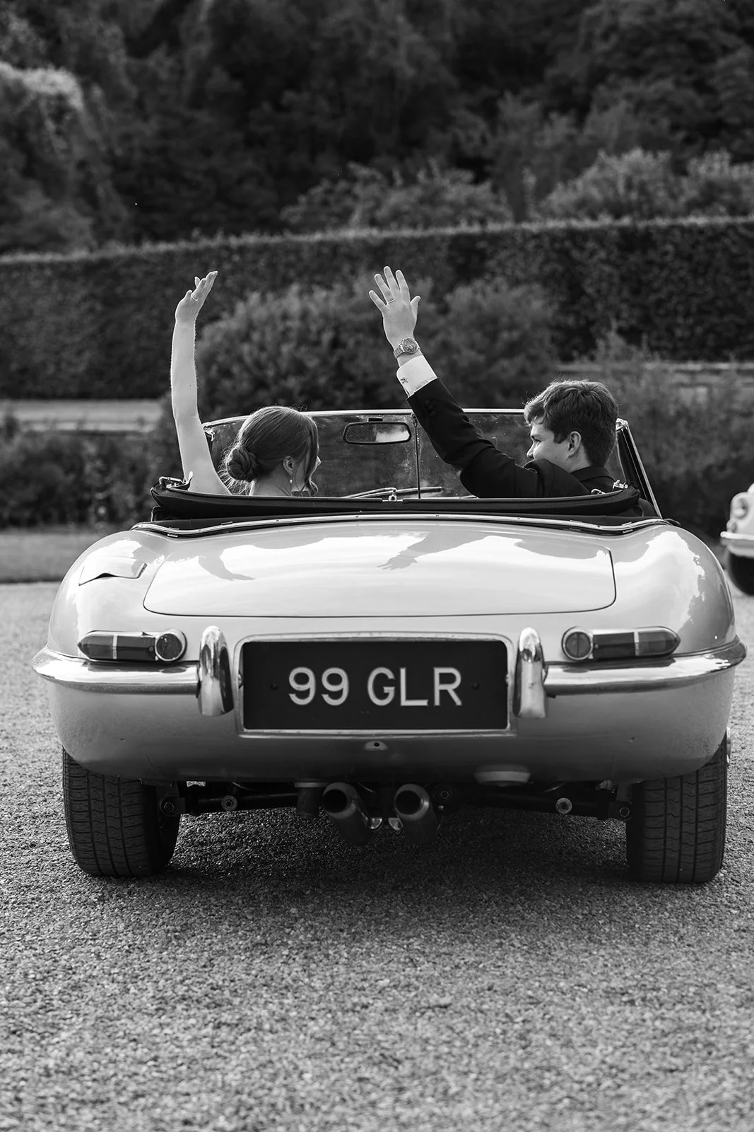 Black and white rear view of a wedding couple waving from a classic convertible car on the driveway at Brympton House in Somerset, photographed by Just James Weddings.