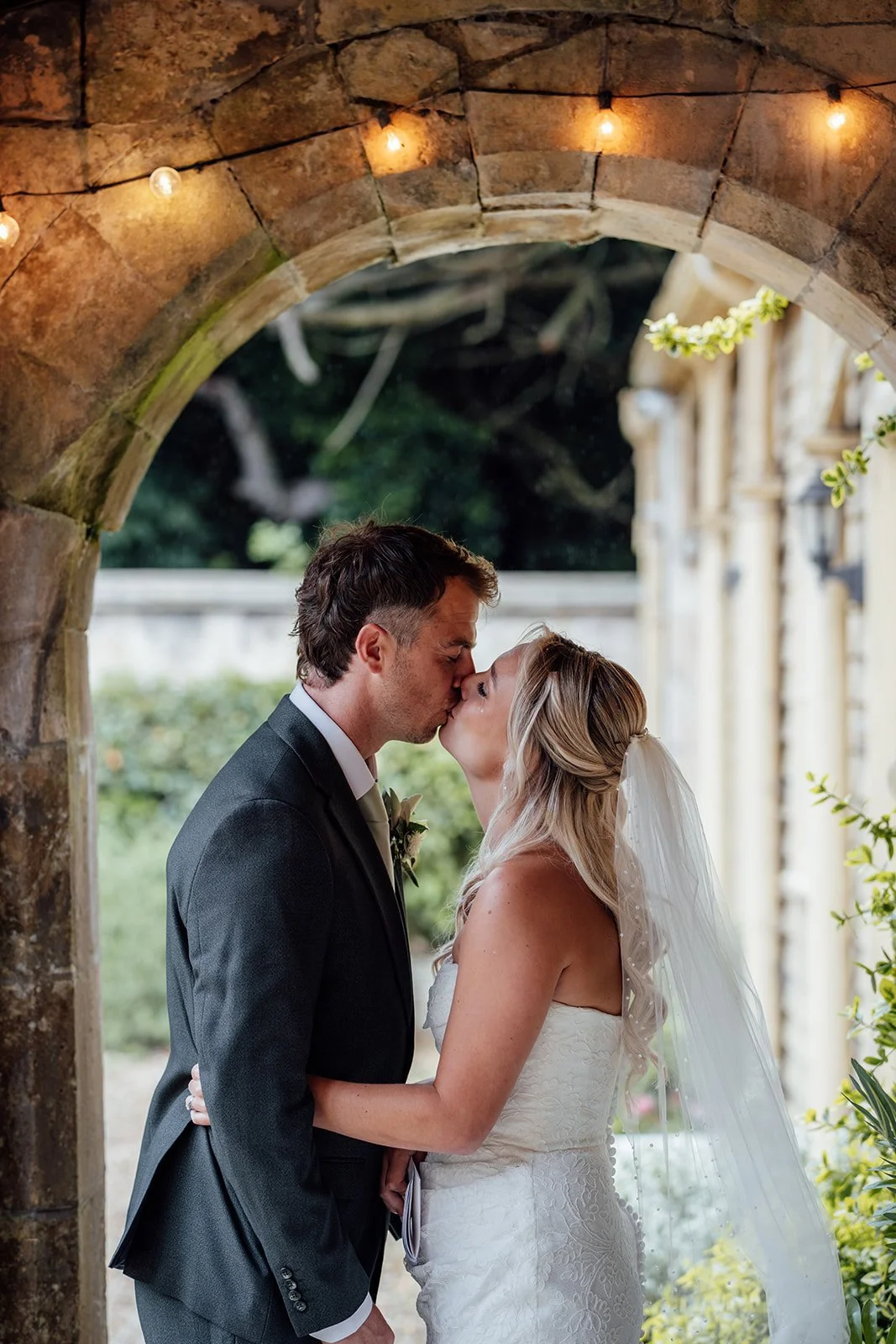 Amy & Matt share a quiet kiss beneath a stone arch at Orchardleigh Estate in Somerset, with warm festoon lights, greenery and historic manor details captured by Just James Weddings.