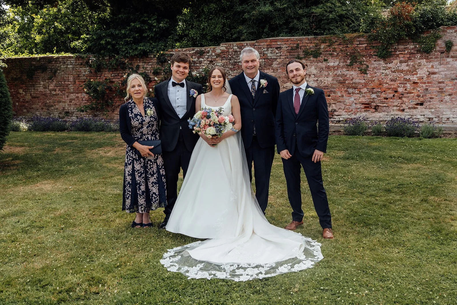 Rachel & Tom pose with three wedding guests in the gardens at Brympton House in Somerset, standing together in front of a weathered red brick wall, photographed by Just James Weddings.