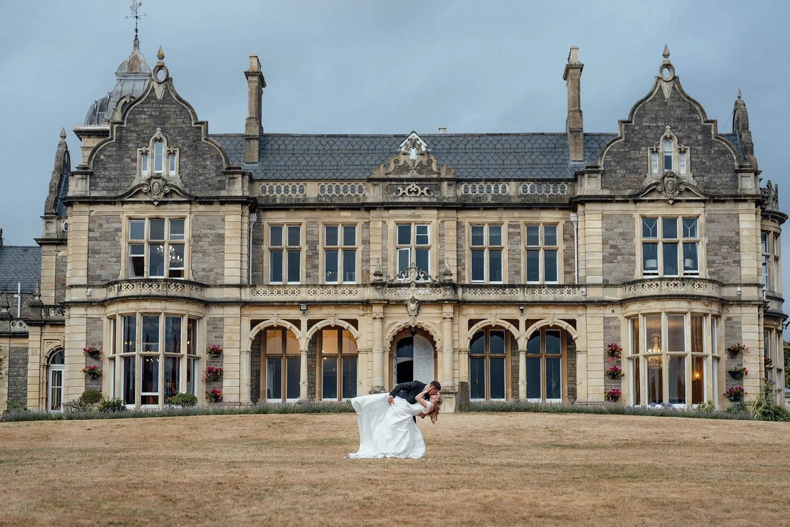 Caitlin & Kevin sharing a playful wedding portrait on the lawn at Clevedon Hall in Somerset, with the historic manor house behind them, photographed by Just James Weddings.