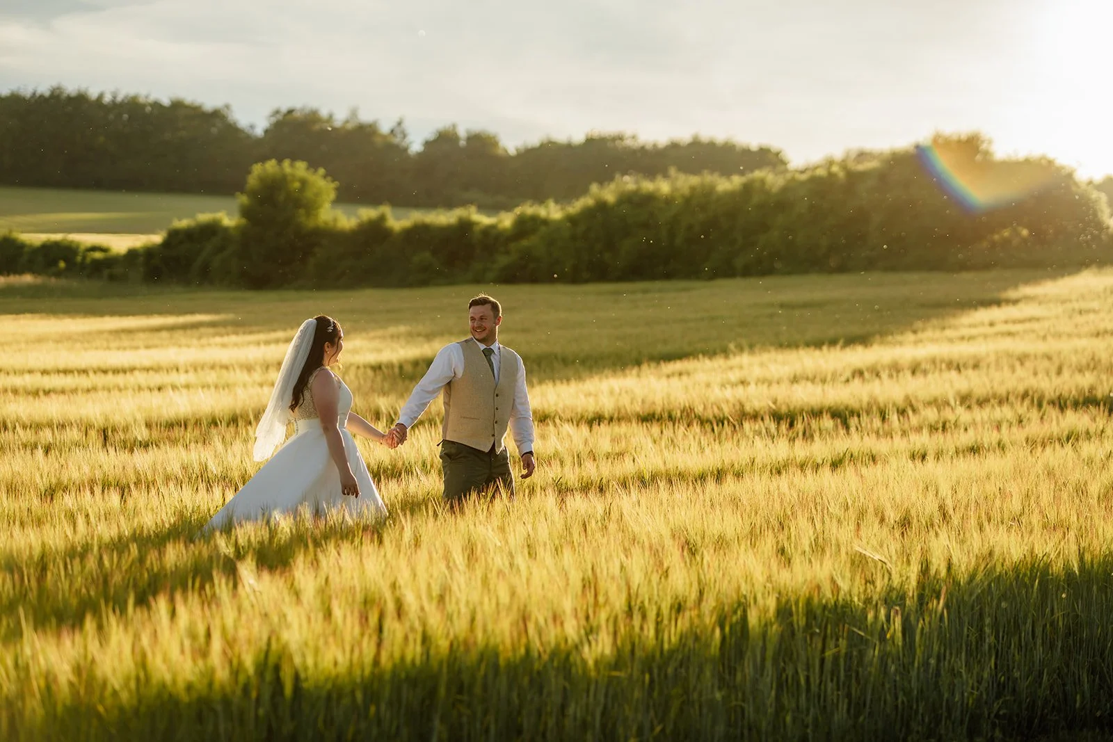 Chloe & Armani walk hand in hand through a sunlit field during golden-hour wedding portraits at The Barn at Upcote in the Cotswolds, photographed by Just James Weddings.