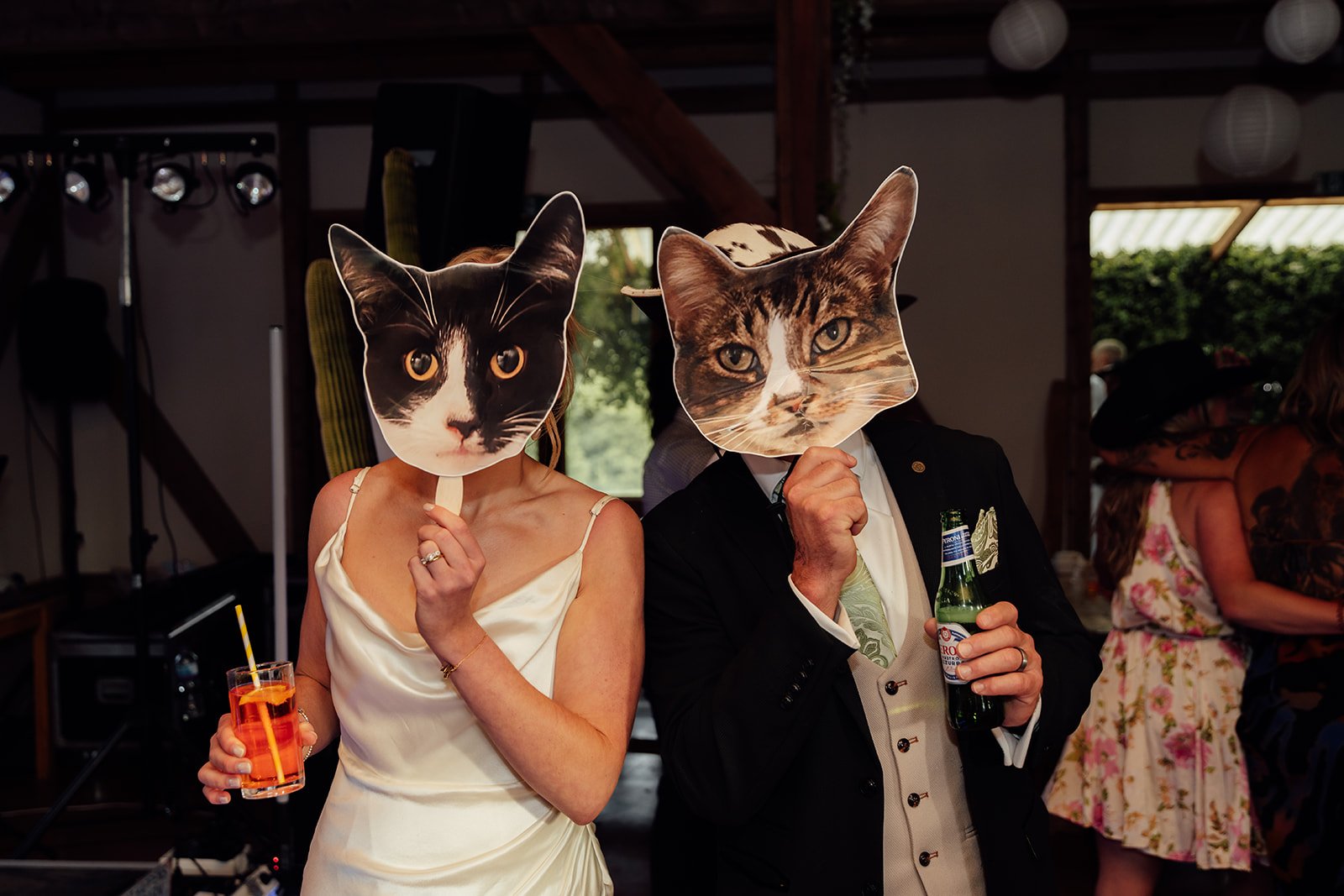 Playful reception photo from Hannah & Ben’s wedding at Grays Barn in Devon, UK, with the couple holding cat face props, drinks and cowboy hats during the evening party, captured with fun, quirky barn wedding energy by Just James Weddings.