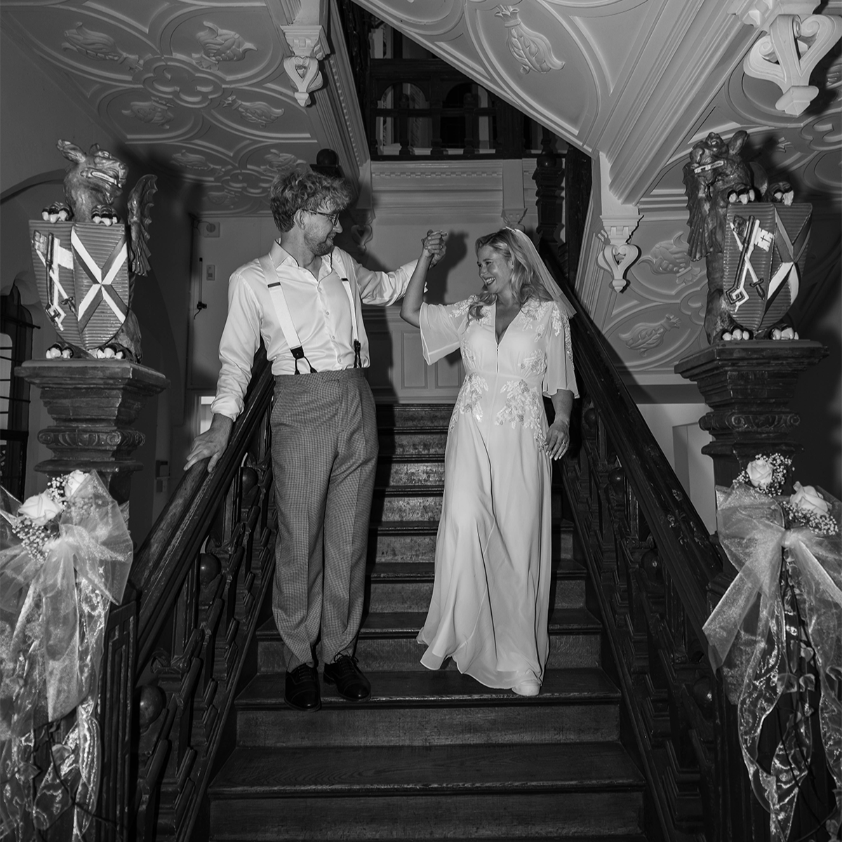 Iona & Will walking down a grand staircase at Bishop’s Palace, somerset, on their wedding day. The bride lifts her dress as the groom guides her, with ornate carved details, stone banisters, and floral arrangements lining the steps.