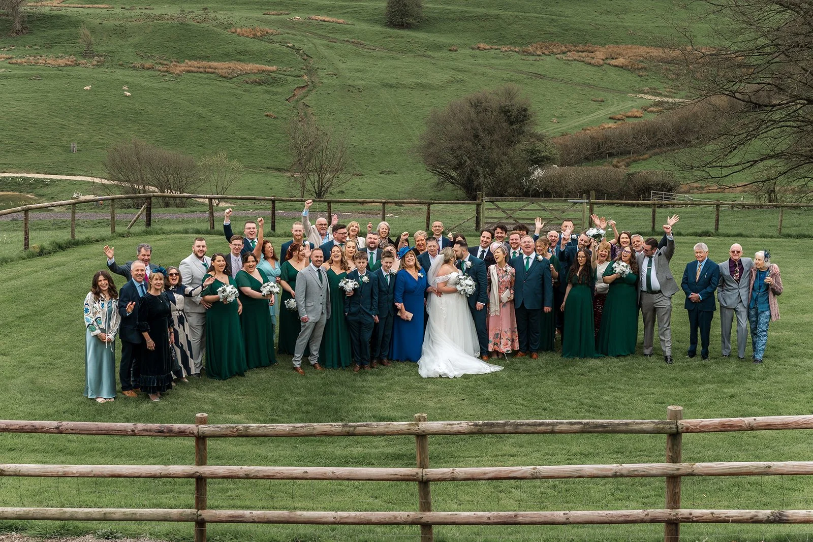 Large group wedding photo of Siobhan & Nicholas with family and guests on the lawn at Kingscote Barn in the Cotswolds, UK, surrounded by rolling countryside views, a joyful rustic barn wedding group portrait captured outdoors after the ceremony.