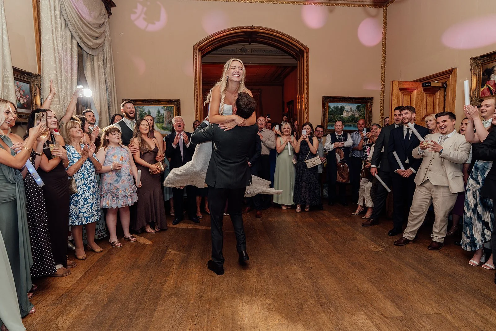 Amy laughs as Matt lifts her during their evening wedding reception at Orchardleigh Estate in Somerset, surrounded by cheering guests on the dance floor, captured by Just James Weddings.