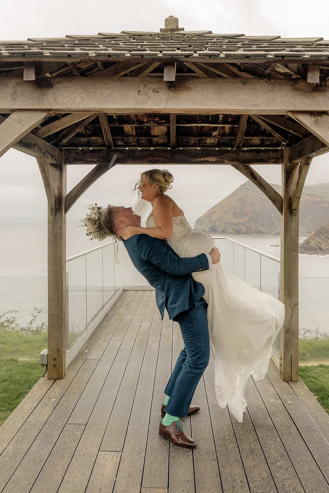 Andrew lifting Daisy in a joyful embrace beneath a wooden gazebo at Sandy Cove Hotel in Devon, with dramatic sea and cliff views behind them, capturing a romantic coastal wedding portrait at this stunning Devon venue.