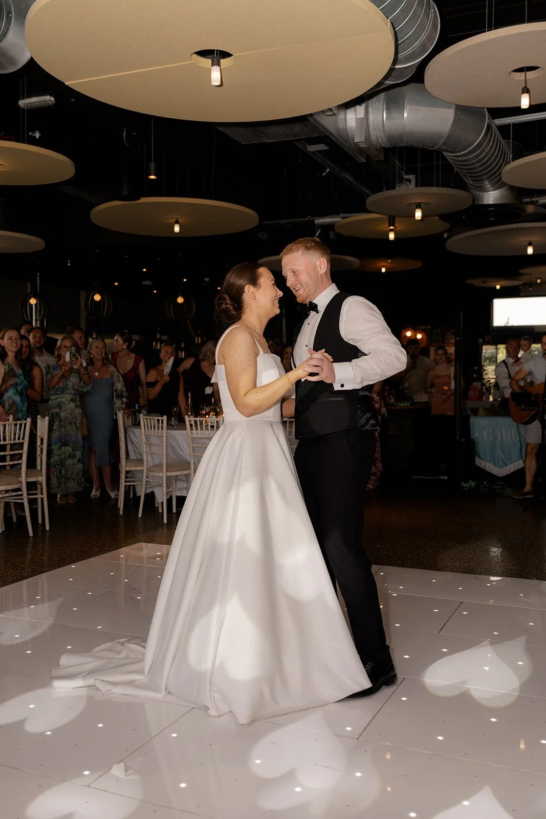 First dance photo of Lydia & Kieran on a white lit dance floor at Hencote Estate in Shropshire, UK, smiling together in the evening reception space with guests watching, modern ceiling lights and soft party lighting around them indoors.