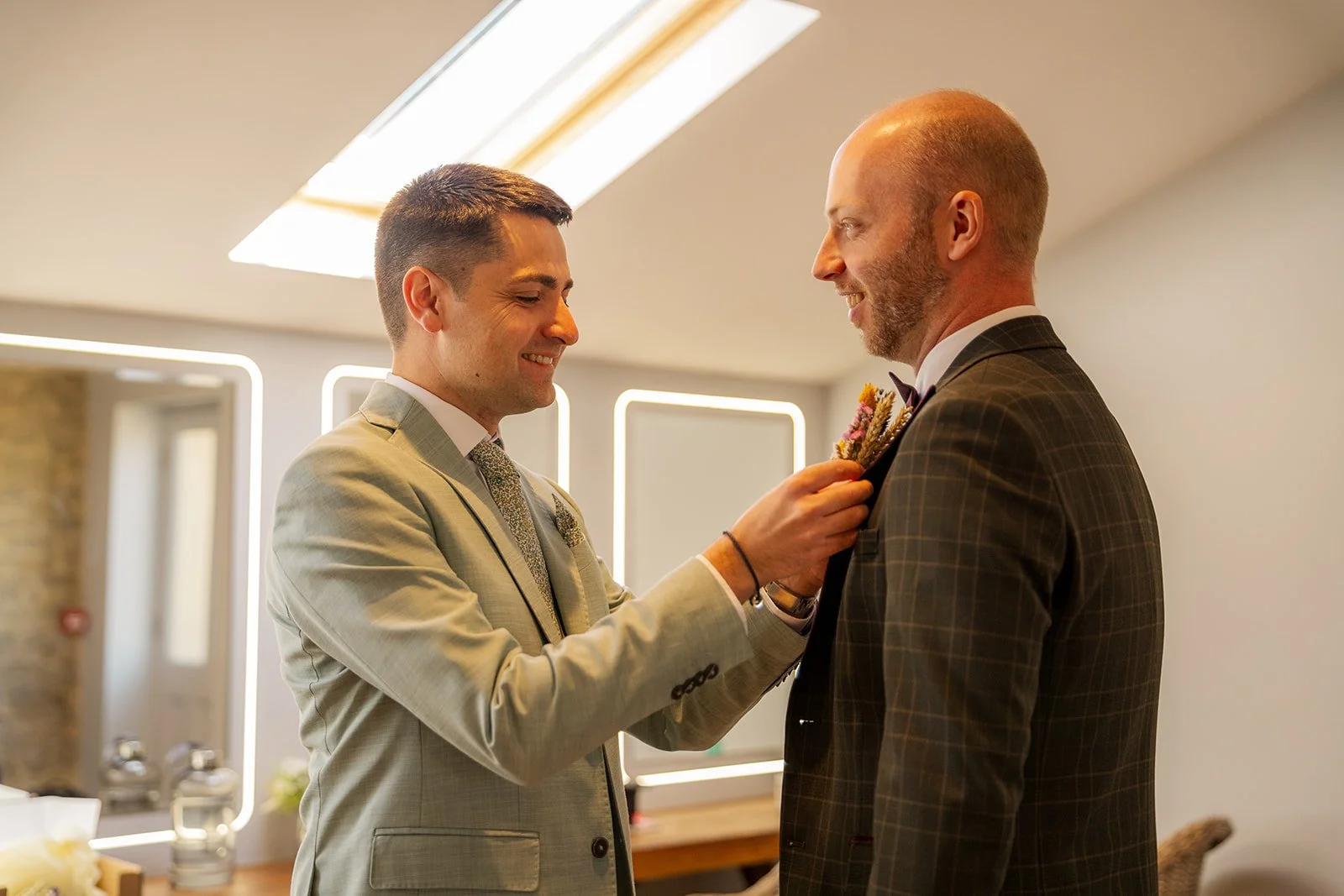 Sam helping Gavin with his buttonhole before the ceremony at The Great Barn at Aynho in the Cotswolds, photographed by Just James Weddings for natural wedding morning moments.
