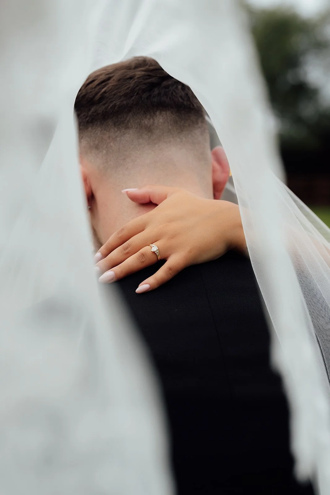 Close-up of Nisha’s engagement ring resting on Jack’s shoulder beneath her flowing veil at Batch Country House in Somerset, UK, creating an intimate wedding portrait focused on their embrace and delicate bridal details by Just James Weddings.