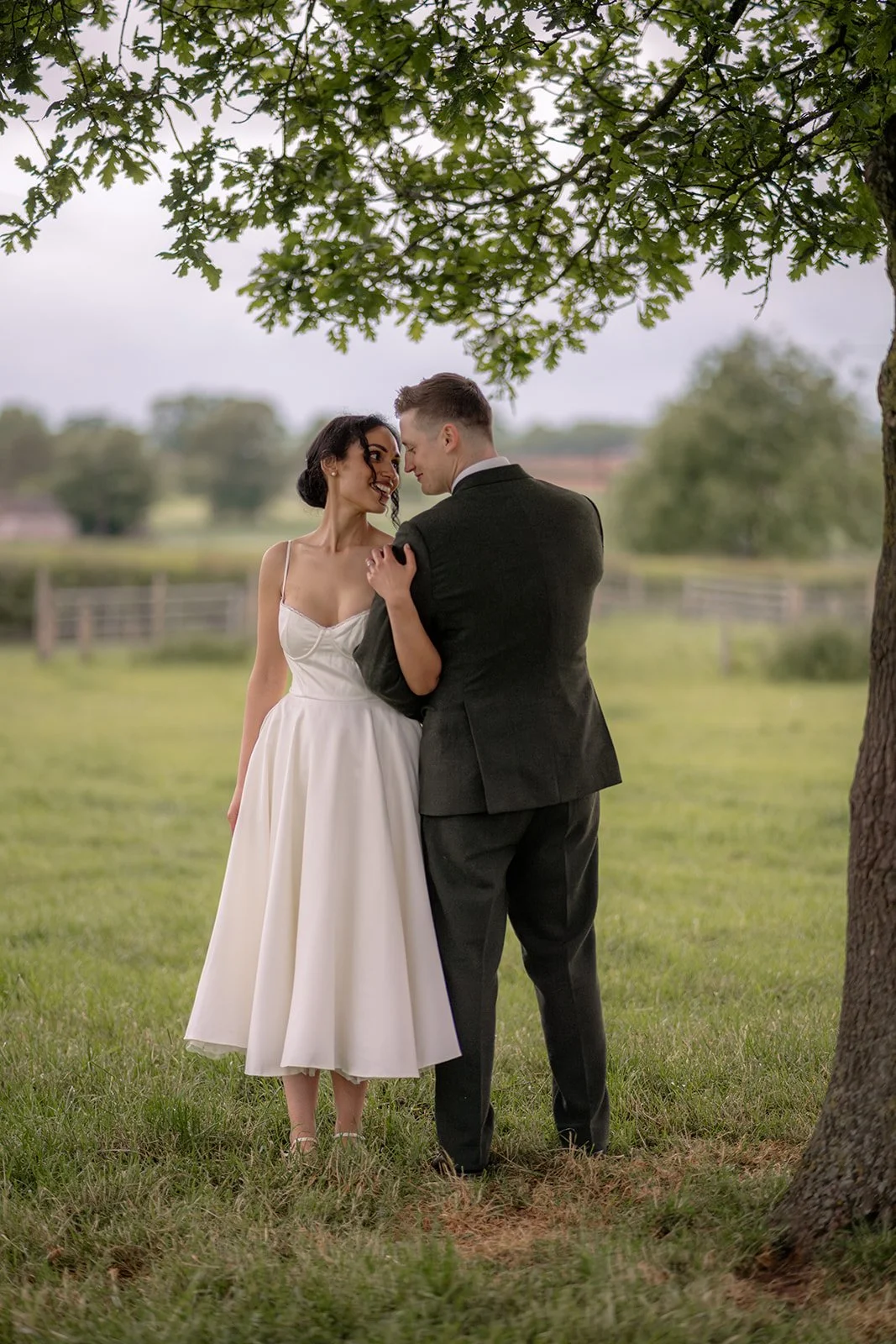 Jaye & Josh stand beneath a leafy tree at Farrington's Farm in Bristol for a relaxed couple portrait, smiling close together in the grass with countryside fields behind them, the bride in a white dress and the groom in a smart green suit nearby.
