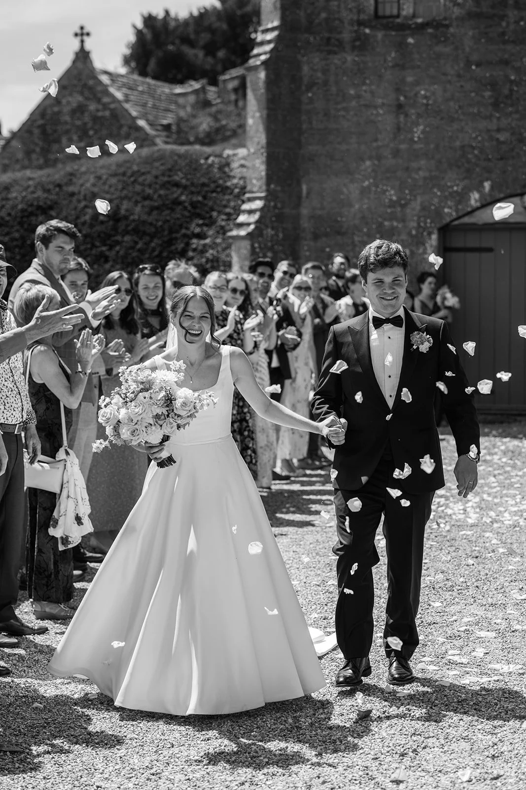 Black and white photo of Rachel & Tom walking hand in hand through falling confetti outside Brympton House in Somerset as wedding guests celebrate, photographed by Just James Weddings.
