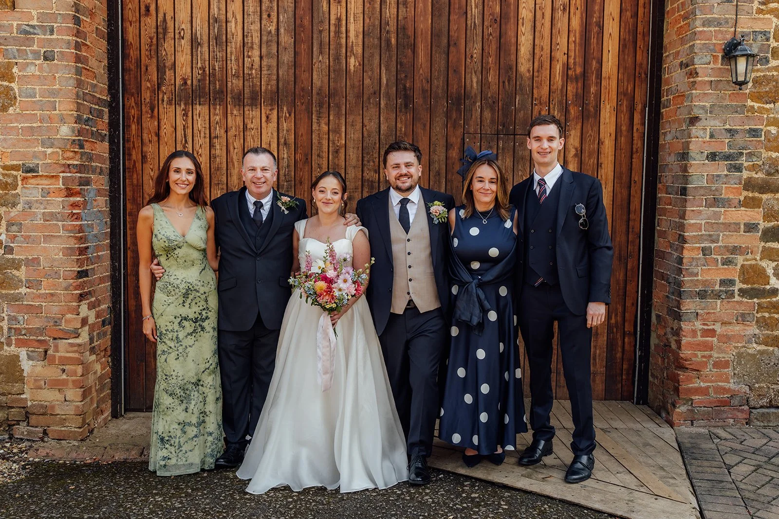 Harriet & Simon stand with close family members for a formal group portrait outside the rustic barn at Dovecote Events in Oxfordshire, photographed by Just James Weddings.