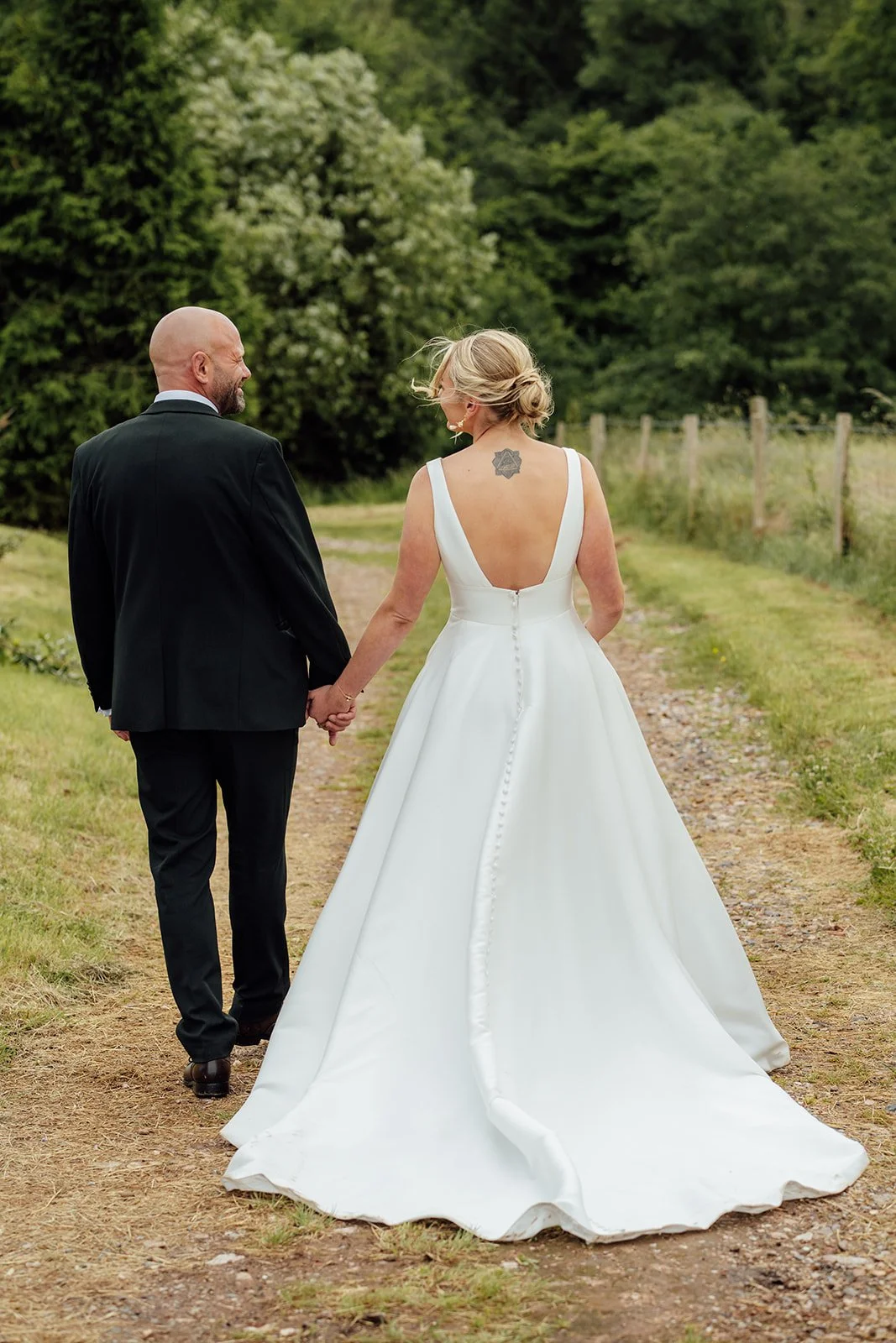 Romantic rear-view portrait of Hannah & Ben walking hand in hand along a countryside path at Grays Barn in Devon, UK, showing Hannah’s flowing white gown, button detail, long train and greenery, photographed naturally by Just James Weddings.