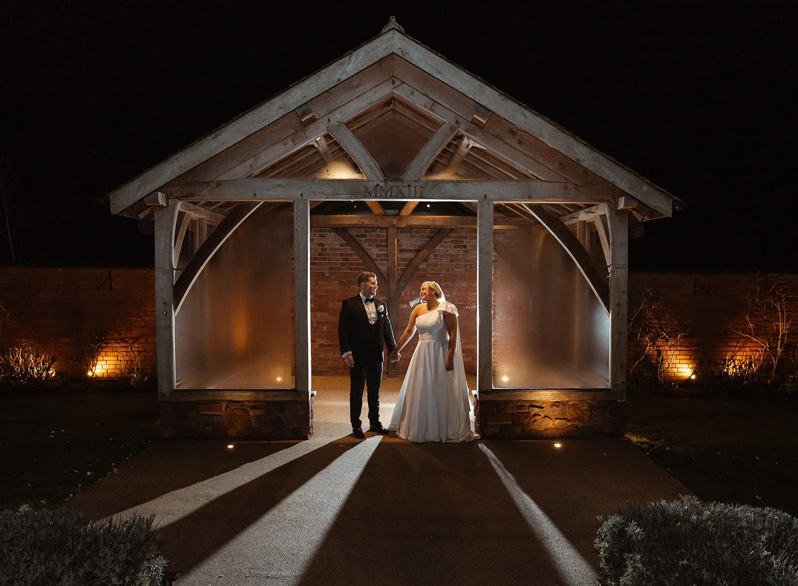 Jessie & Jay stand hand in hand for a night wedding portrait at Upton Barn & Walled Garden, Devon, framed by a timber pavilion with warm uplighting and dramatic shadows, capturing a romantic evening moment at their countryside wedding venue.