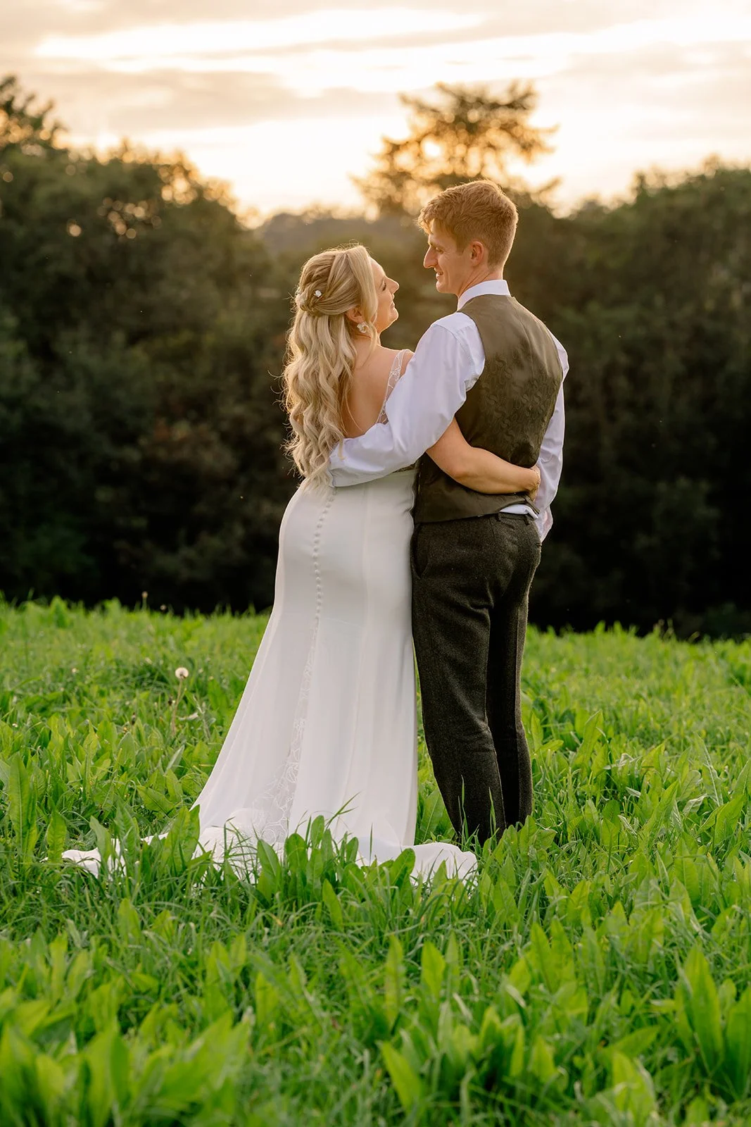 Abi & Josh standing together in a green field at The Cider Barn in Somerset, with the bride and groom wrapped in each other’s arms at sunset, photographed by Just James Weddings for natural countryside wedding portraits.