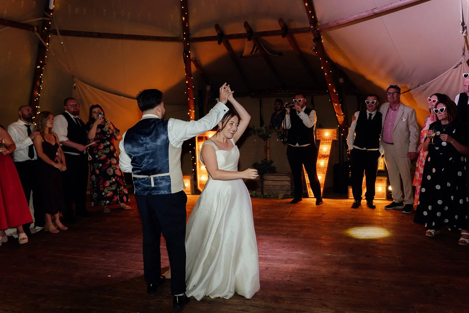 Harriet & Simon dance together inside the tipi as wedding guests gather around them beneath warm fairy lights at Dovecote Events in Oxfordshire, photographed by Just James Weddings.