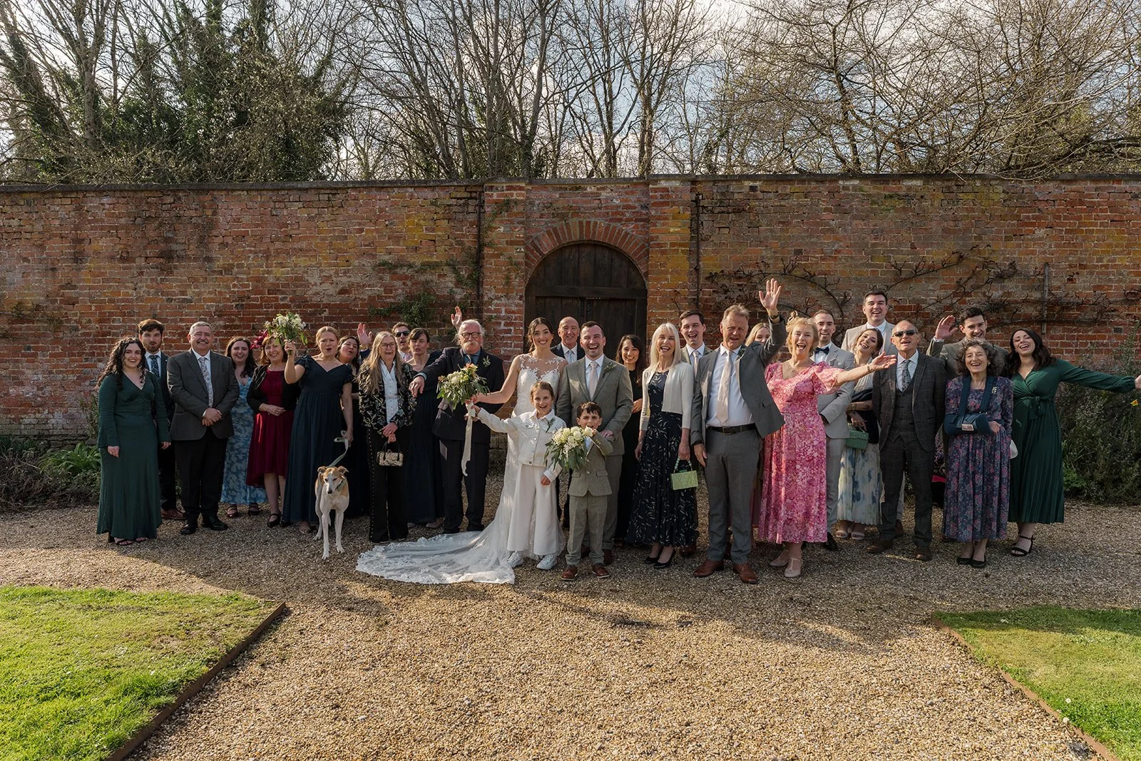 Anna & Chris stand with their wedding guests outside Elmhay Park in Somerset, UK, smiling in front of a long brick garden wall as family and friends wave to camera. A relaxed group portrait from Anna & Chris’s wedding day in the spring sunshine.