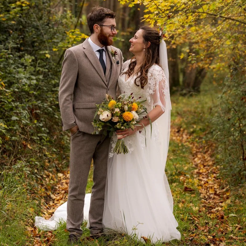 Maisie & Andrew standing together on a leafy woodland path at Stockbridge Farm Barn, dorset, on their wedding day. The bride holds a bouquet of yellow and white flowers as the groom smiles at her, with autumn leaves around them.