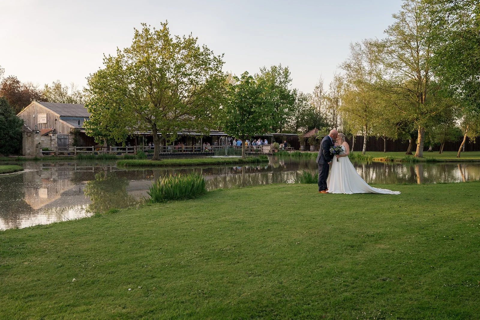 Debbie and Nigel share a golden hour kiss beside the lake at Yarlington Barn in Somerset, with the rustic barn, guests, trees and water reflections framing a peaceful countryside wedding portrait in the venue gardens.