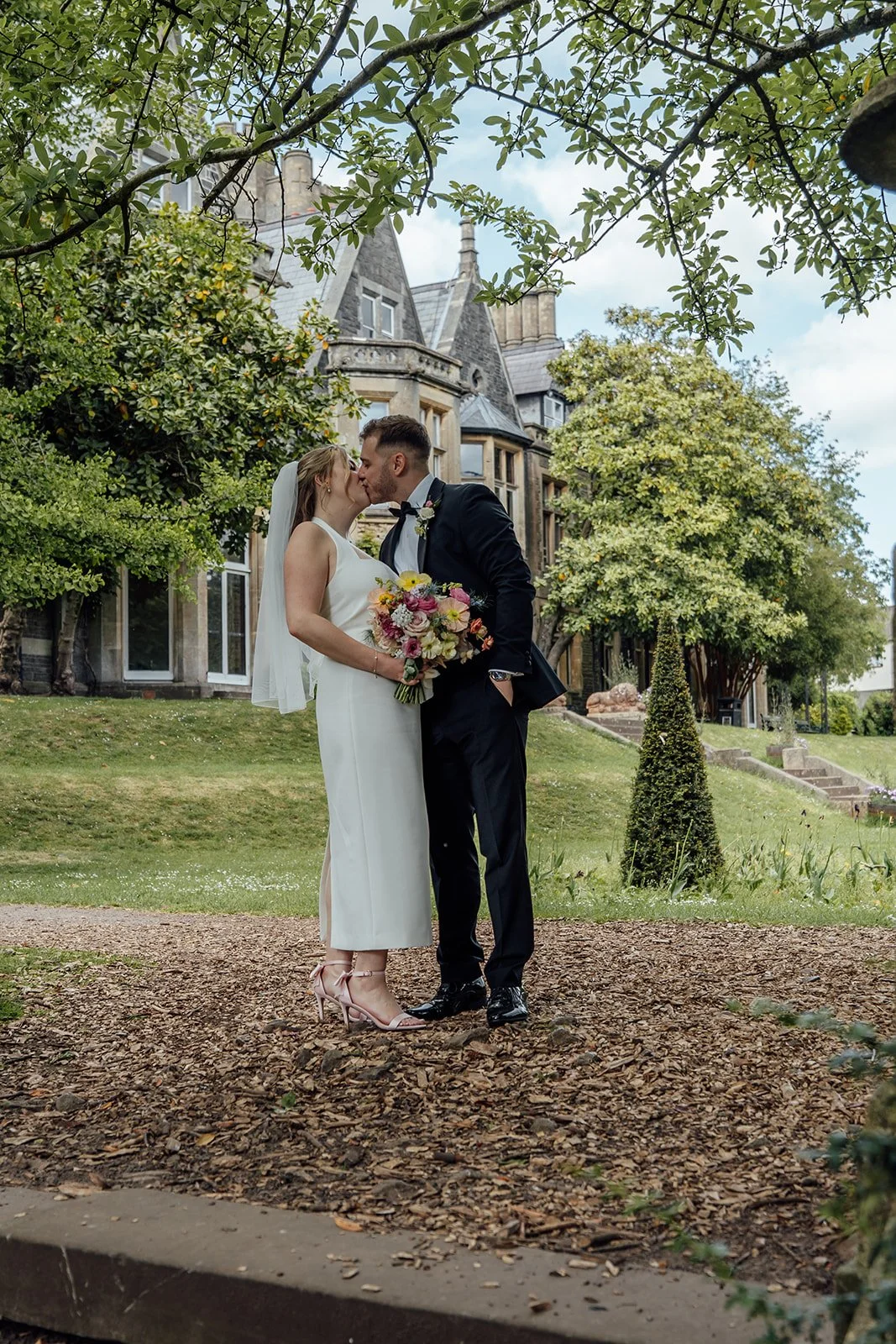 Bryony & Max kiss beneath leafy branches in the gardens at Insole Court in Cardiff, with the historic house behind them, Bryony holding her colourful bouquet and the couple framed by greenery for a relaxed outdoor wedding portrait on the lawn.