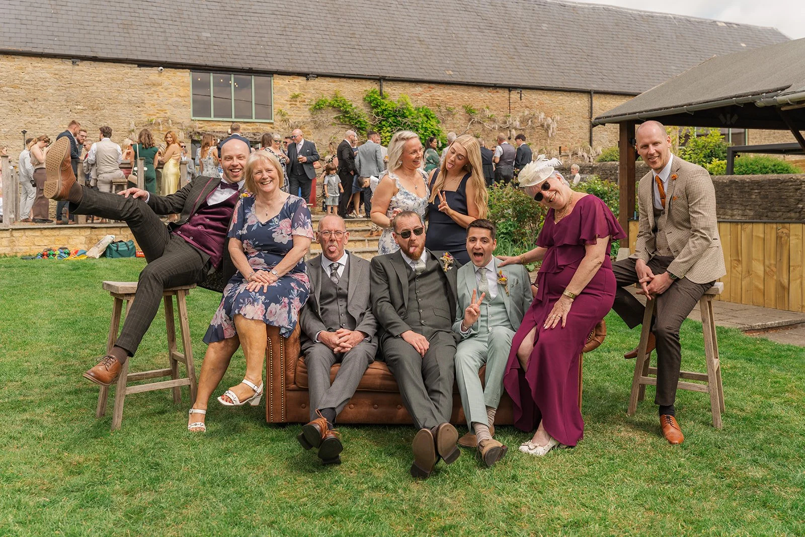 Sam & Gavin posing with guests on vintage sofas at The Great Barn at Aynho in the Cotswolds, photographed by Just James Weddings for fun relaxed wedding group photos.