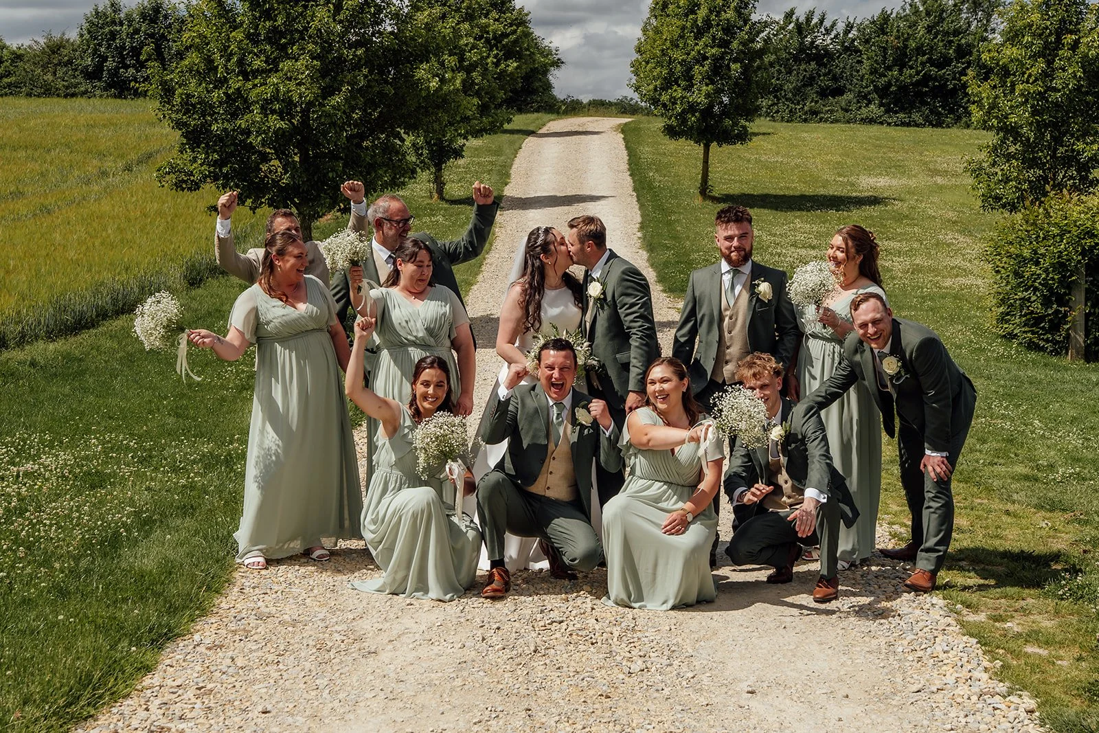 Chloe and Armani kiss while their bridesmaids and groomsmen celebrate around them on the tree-lined drive at The Barn at Upcote in Gloucestershire, photographed by Just James Weddings.