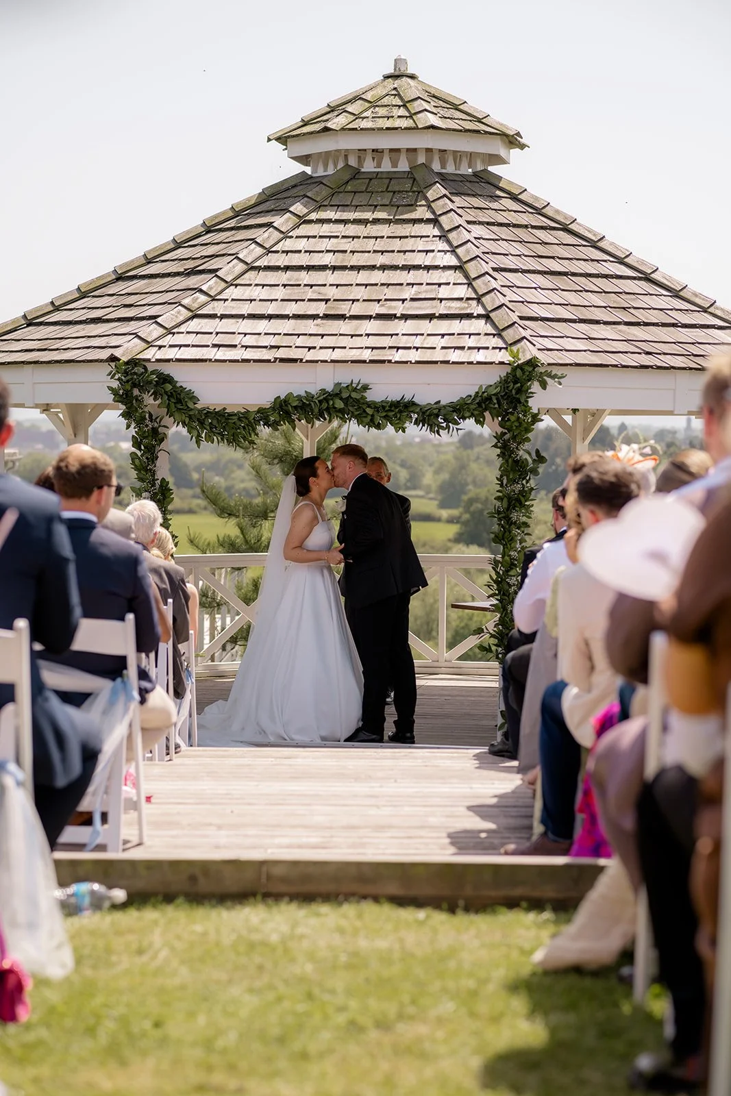 Outdoor ceremony photo of Lydia & Kieran sharing their first kiss beneath a white gazebo at Hencote Estate in Shropshire, UK, with guests watching from the aisle and sunny countryside views behind them during the wedding ceremony in summer.