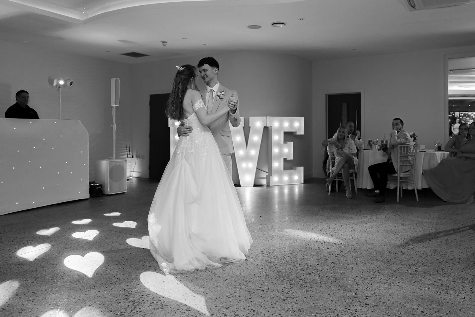 Hannah & Cameron share their first dance beneath glowing LOVE letters during the evening reception at Sandy Cove Hotel, North Devon. Heart-shaped lights reflect on the floor as the couple hold each other during this romantic moment.