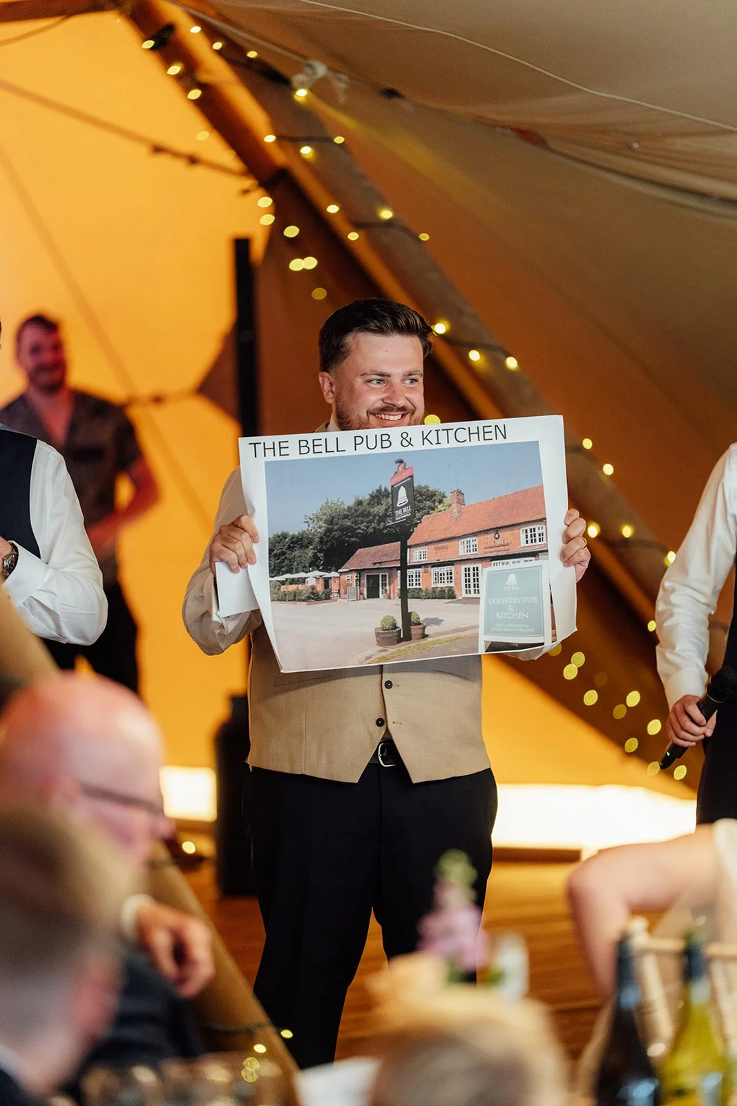 Simon smiles while holding up a large photo of The Bell Pub & Kitchen during a wedding speech inside the tipi at Dovecote Events in Oxfordshire, photographed by Just James Weddings.