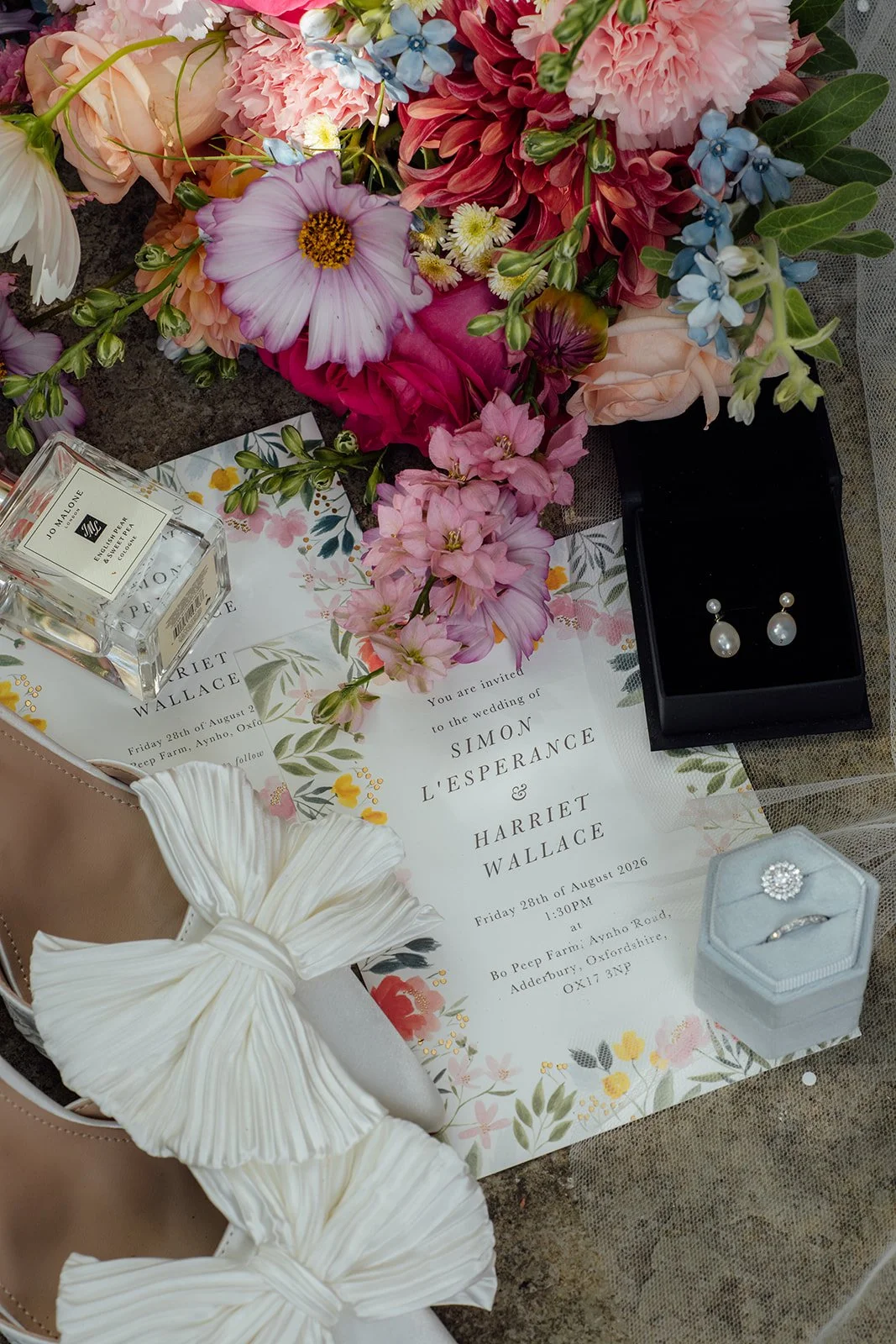 Flat lay of Harriet’s colourful bouquet, floral wedding invitation, pearl earrings, perfume, rings and ivory bow shoes at Dovecote Events in Oxfordshire, photographed by Just James Weddings.