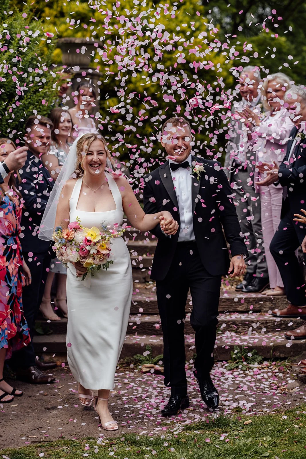 Bryony & Max walk hand in hand through pink and white confetti outside Insole Court in Cardiff, smiling as guests cheer around them, with Bryony holding her colourful bouquet during a joyful garden wedding celebration in the grounds nearby.