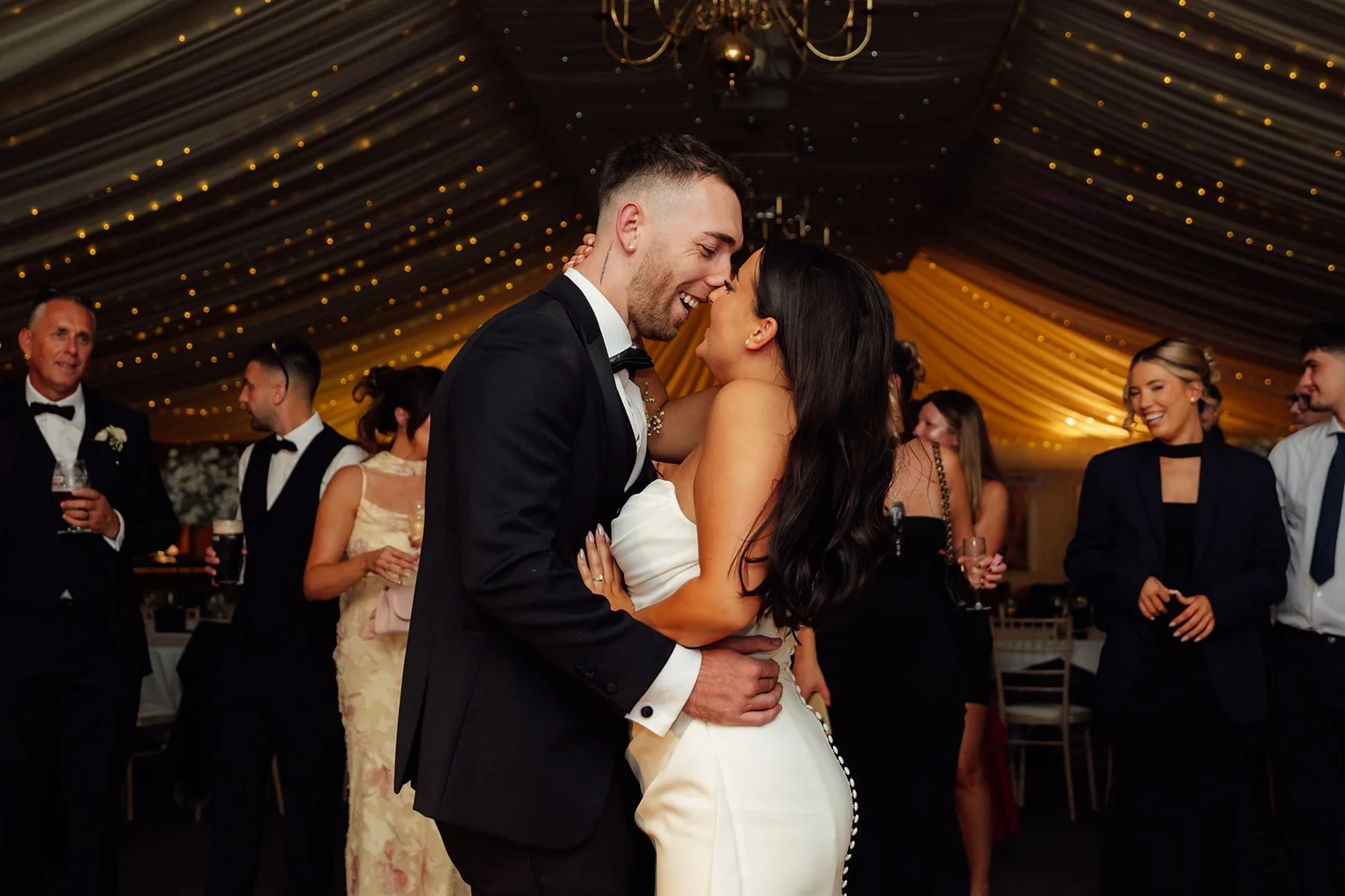 Nisha and Jack share a joyful moment on the dance floor during their wedding reception at Batch Country House in Somerset, UK, surrounded by smiling guests beneath warm draping and fairy lights, photographed naturally by Just James Weddings.