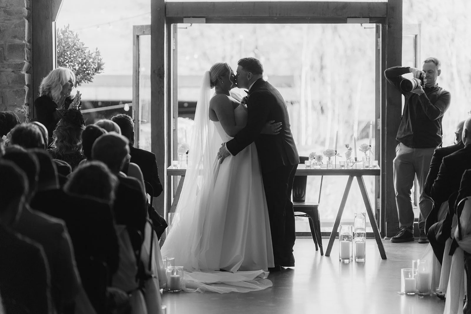 Jessie & Jay share their first kiss at Upton Barn & Walled Garden in Devon, UK, during an indoor wedding ceremony, surrounded by guests, soft candlelight, and a bright doorway backdrop that frames the bride and groom in a timeless moment.