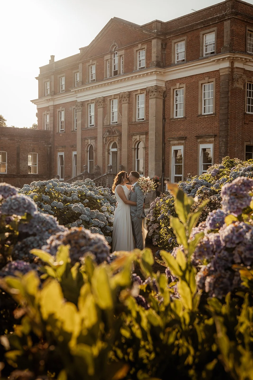 Michelle and Andy share a kiss among blue hydrangeas in the gardens of Crowcombe Court in Somerset, UK, during warm golden-hour wedding portraits.