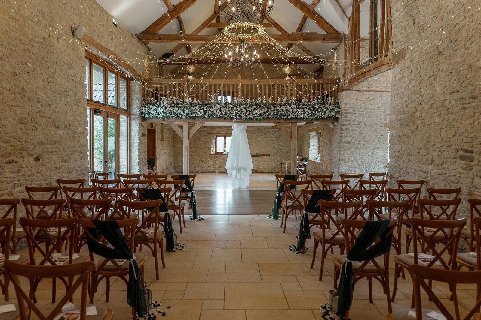 Wide view of the ceremony room at Kingscote Barn in the Cotswolds, UK, with rows of wooden chairs, fairy lights and Siobhan & Nicholas' wedding dress hanging beneath floral arrangements for a rustic barn wedding ceremony set-up before guests.