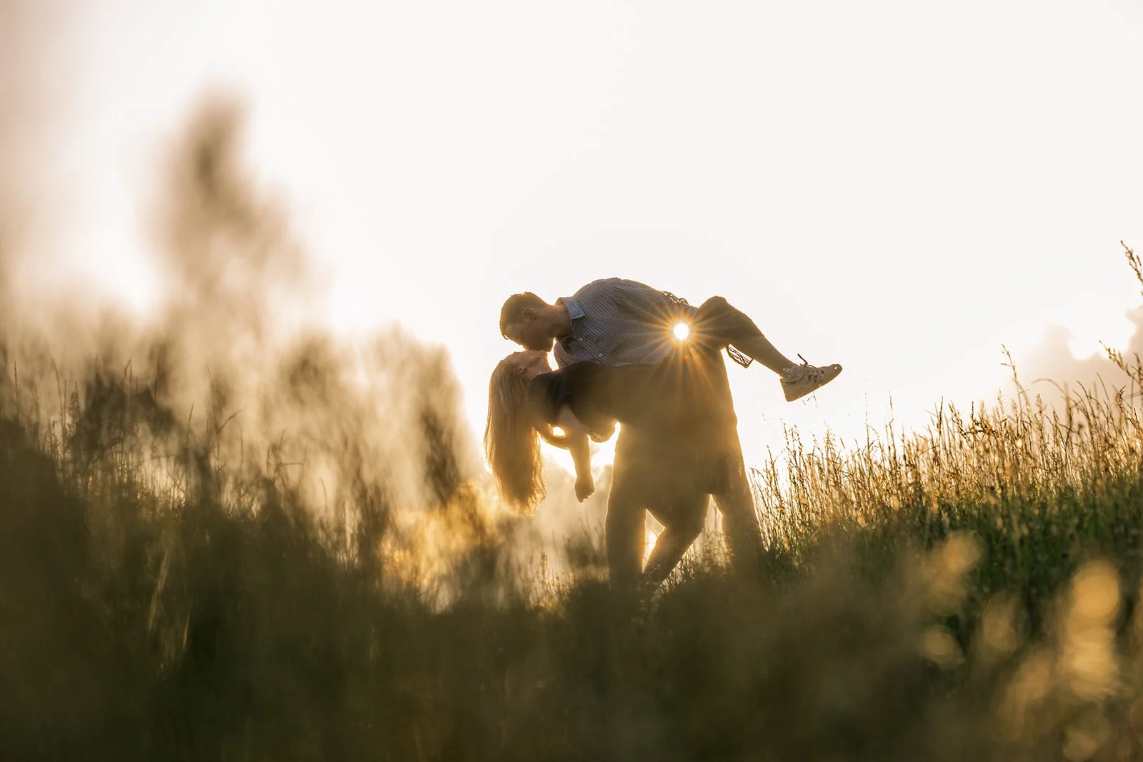 Caitlin and Kevin sharing a sweet kiss as Kevin lifts her in a golden Somerset field in the evening, surrounded by long grass and warm sunset light during their relaxed countryside engagement photoshoot captured by Just James Weddings outdoors.