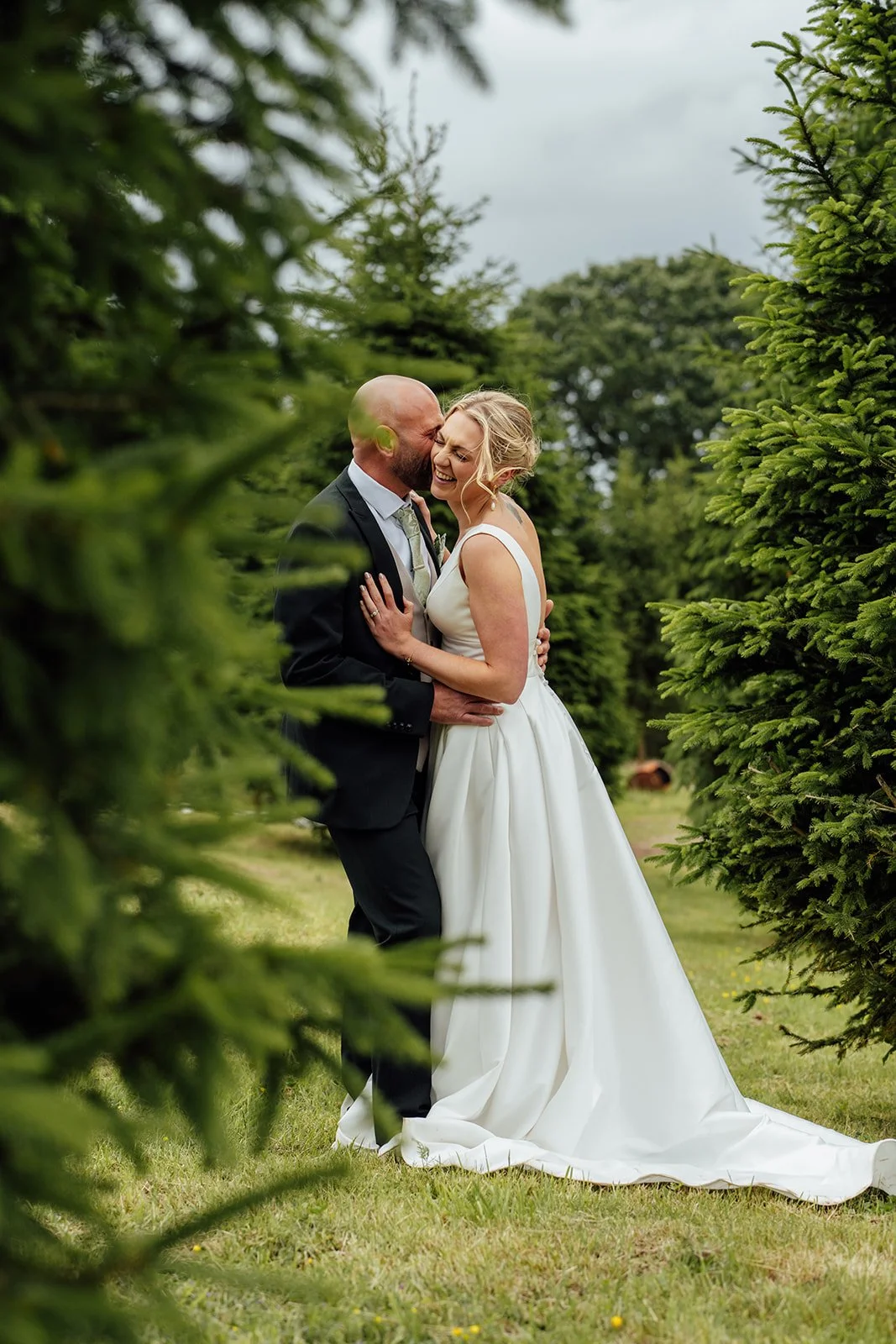 Joyful couple portrait of Hannah & Ben at Grays Barn in Devon, UK, laughing together among evergreen trees after the ceremony, with her white gown and his dark suit framed by soft countryside greenery, photographed warmly by Just James Weddings.