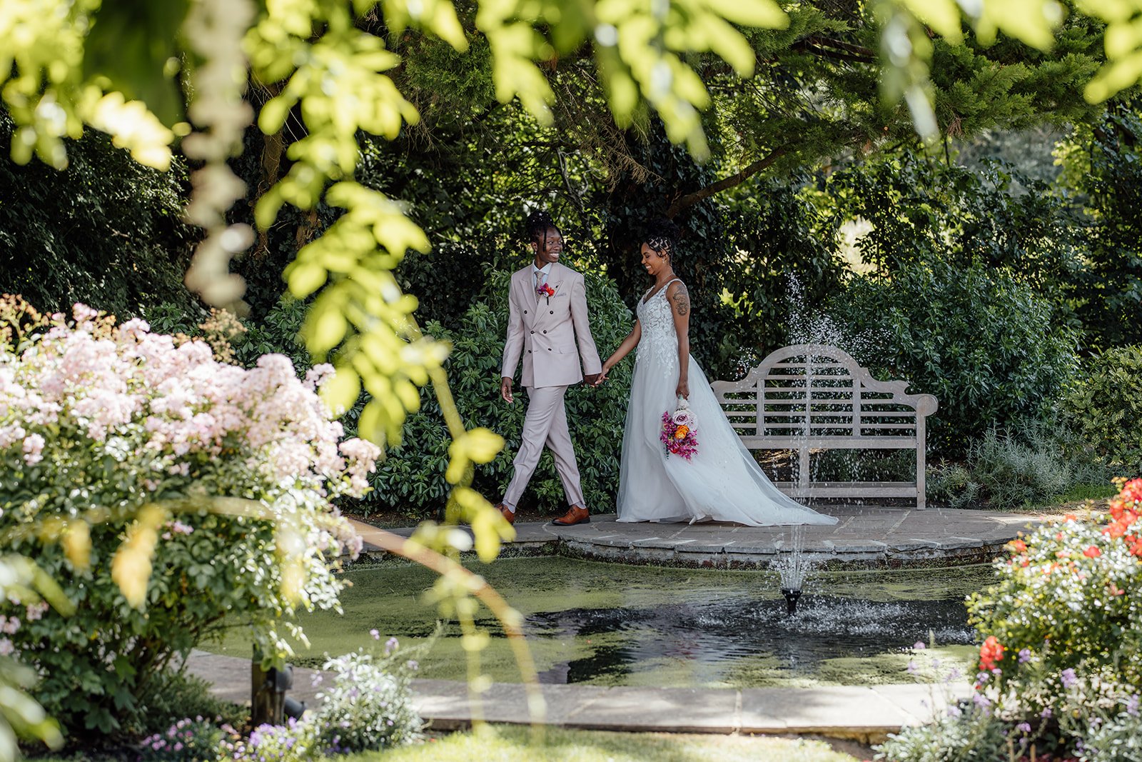 Kasey and Larisha walking hand in hand beside the garden pond and fountain during their wedding portraits at Oaks Farm Weddings in Surrey.