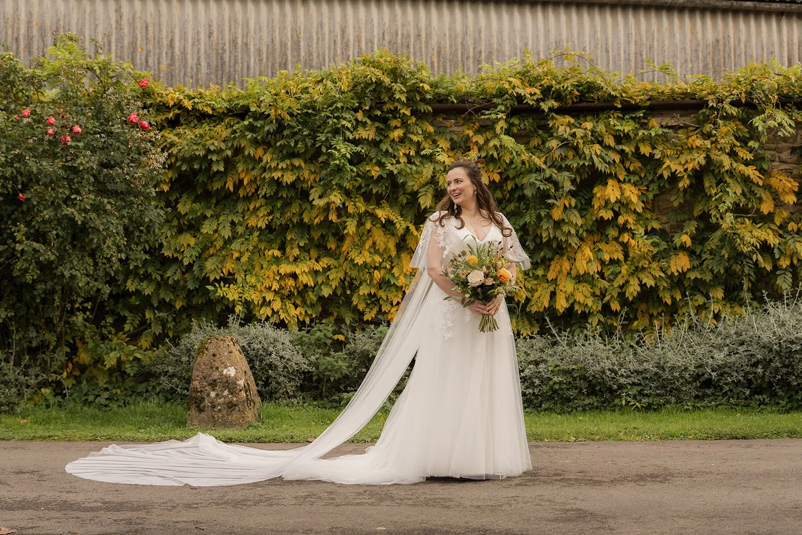 Maisie standing with her autumn bouquet outside Stockbridge Farm Barn in Dorset, surrounded by seasonal greenery and warm autumn colours, photographed by Just James Weddings.