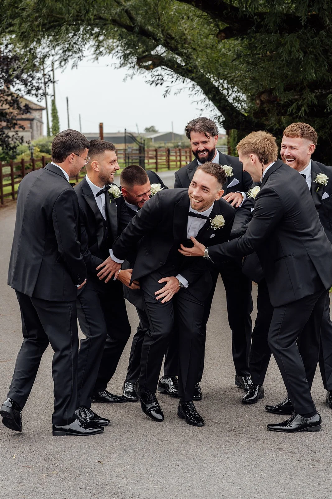Jack laughs with his groomsmen in matching black suits outside Batch Country House in Somerset, UK, as the group crowd around him for a relaxed and playful wedding party portrait full of personality, photographed by Just James Weddings.